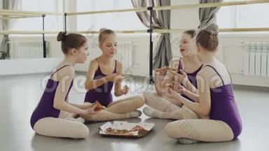 穿着亮色紧身衣的快乐女孩正一起坐在芭蕾舞工作室的地板上吃披萨和聊天。 美味的食物